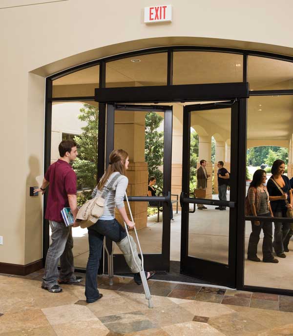a boy presses a button for an automatic door operator so a girl on crutches can walk outside